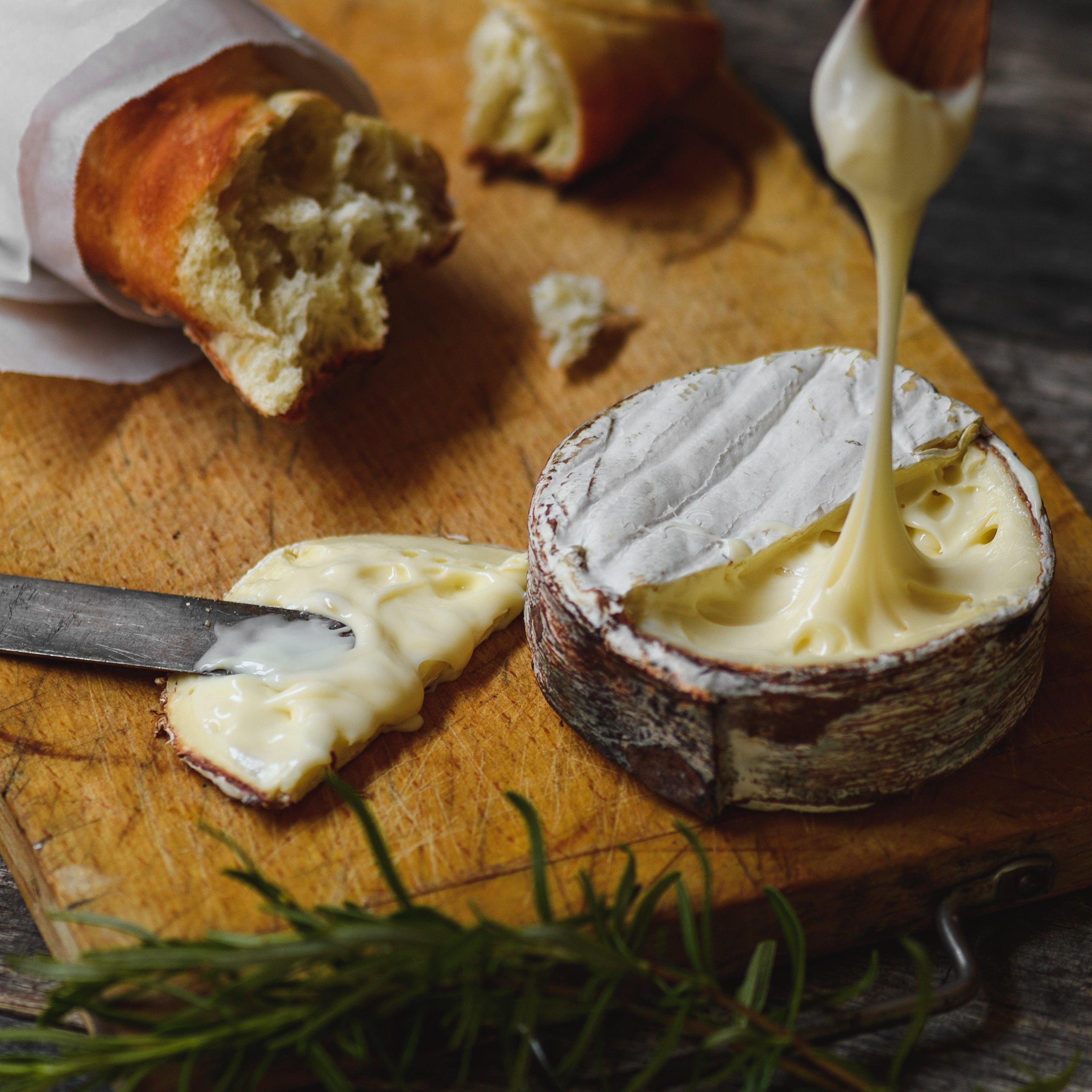 A wheel of soft white cheese with a rind, shown on a wooden cutting board with bread, a knife, and a spoon pouring melted cheese over it. Rosemary is visible in the foreground on a rustic wooden table.