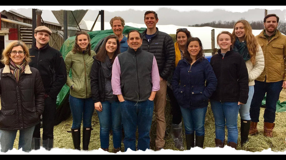 Group of people standing outdoors in winter clothing with a barn and snow in the background.
